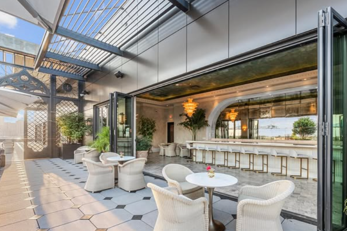 Aperitivo Houston patio with white wicker chairs, round tables, open-air bar, gold chandeliers, potted plants, and modern design under a blue sky.