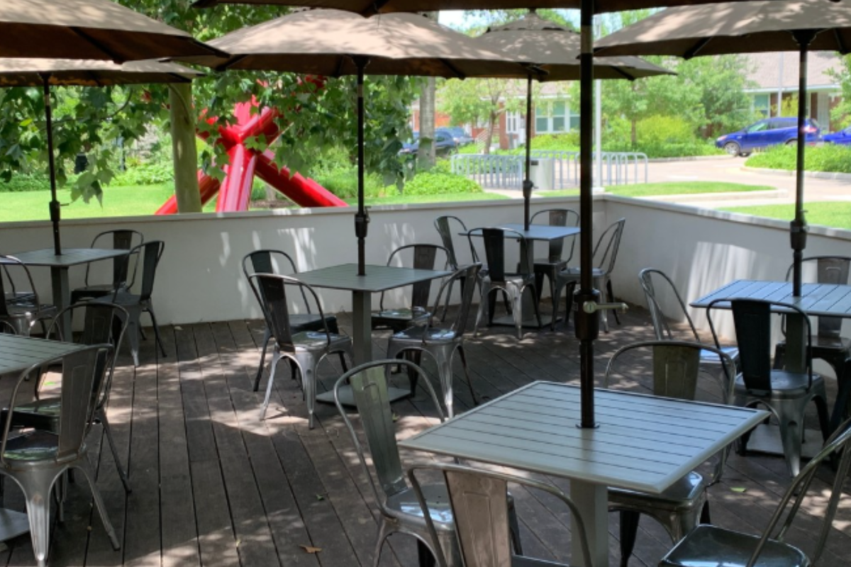 Outdoor patio at Bistro Menil with metal chairs, square tables, beige umbrellas, wood deck flooring, and a red abstract sculpture in the garden view.