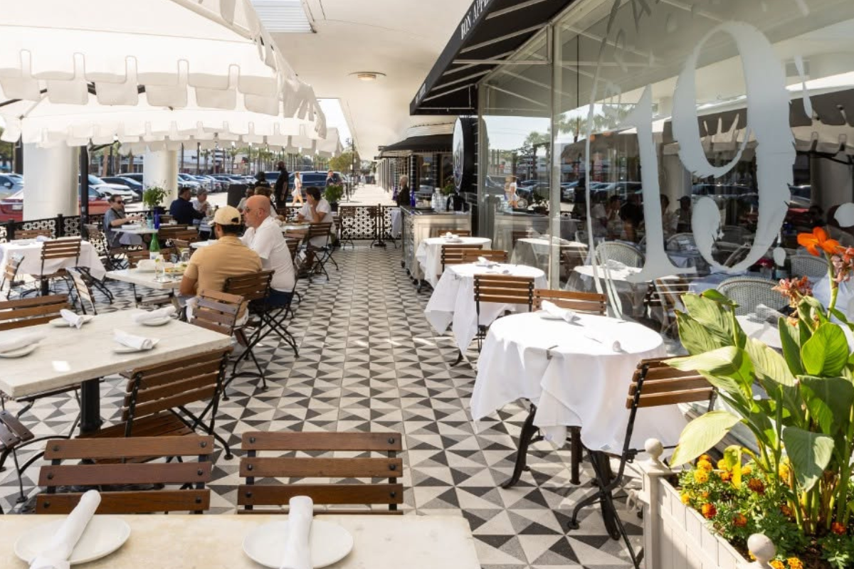 Brasserie 19 outdoor patio with white umbrellas, wooden tables, black and white tile flooring, diners enjoying meals, and potted flowers by the entrance.