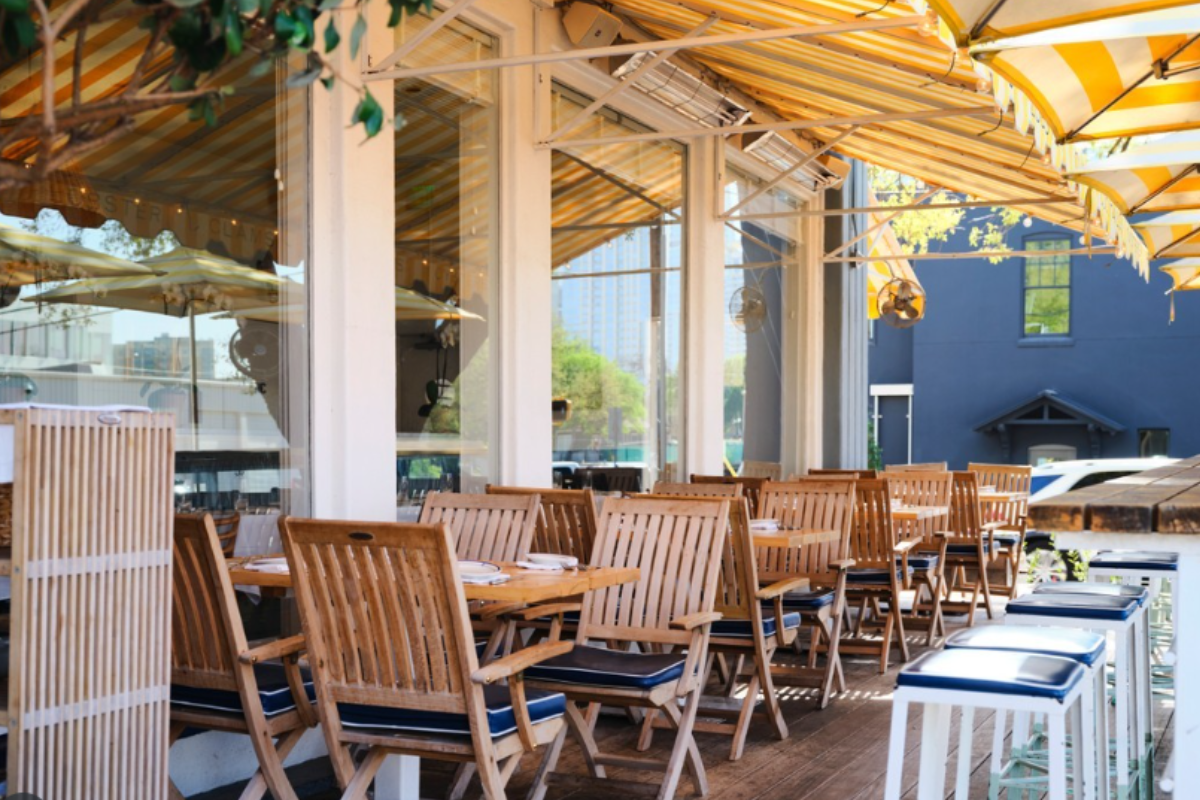 Clark’s Oyster Bar patio with wooden tables, chairs, striped yellow awning, blue cushioned stools, and sunny outdoor seating with city views.