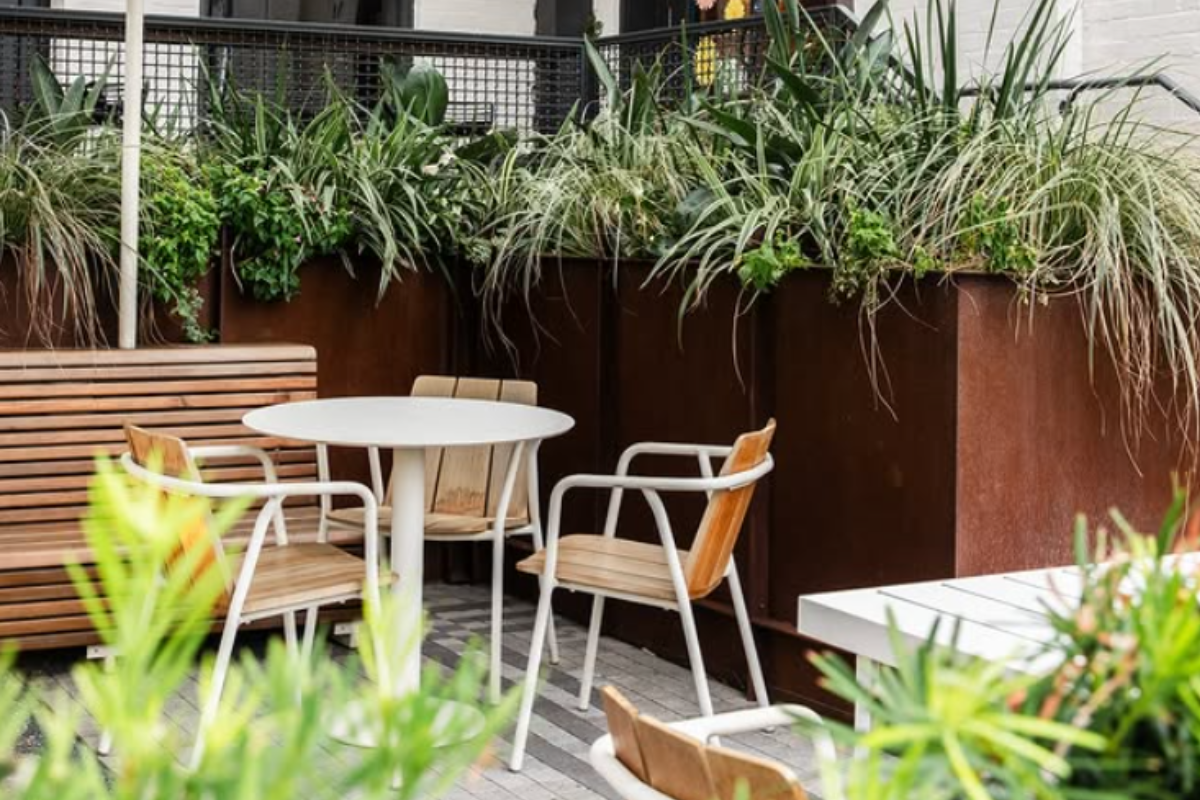 Dandelion Cafe patio with white round table, wooden chairs, bench seating, tall planters filled with green plants, and cozy outdoor garden vibe.