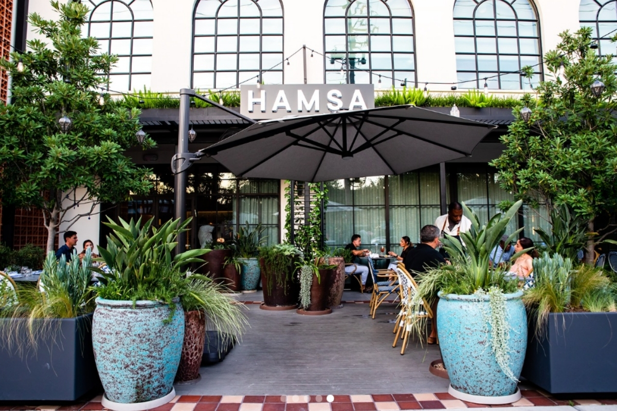Hamsa restaurant patio with large potted plants, black umbrella, string lights, outdoor seating, and guests dining in front of arched windows.