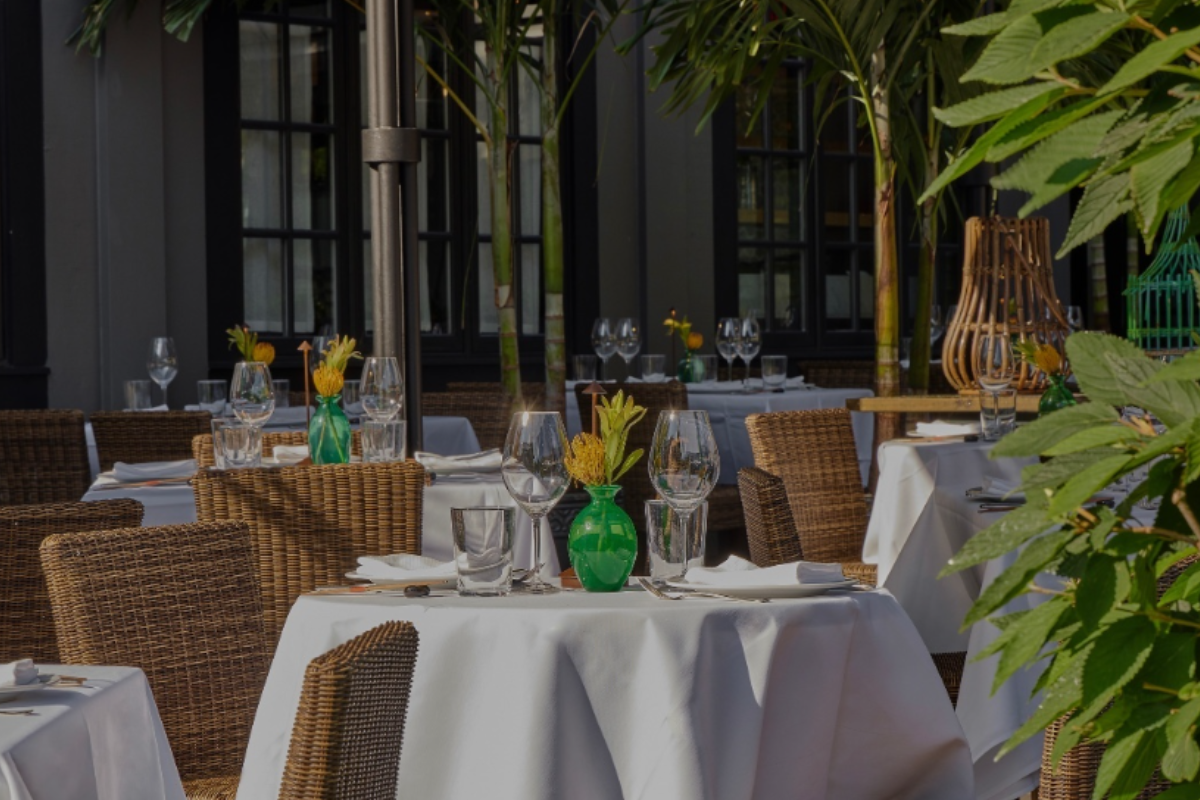 Le Colonial patio with wicker chairs, round tables with white linens, glassware, green vases with flowers, tropical plants, and elegant outdoor setting.