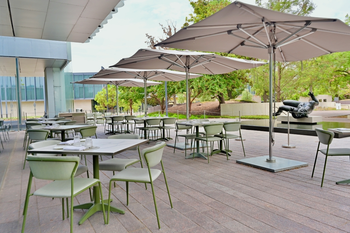 Le Jardinier outdoor patio with green chairs, modern tables, large gray umbrellas, wood deck, garden views, and black sculpture in the background.