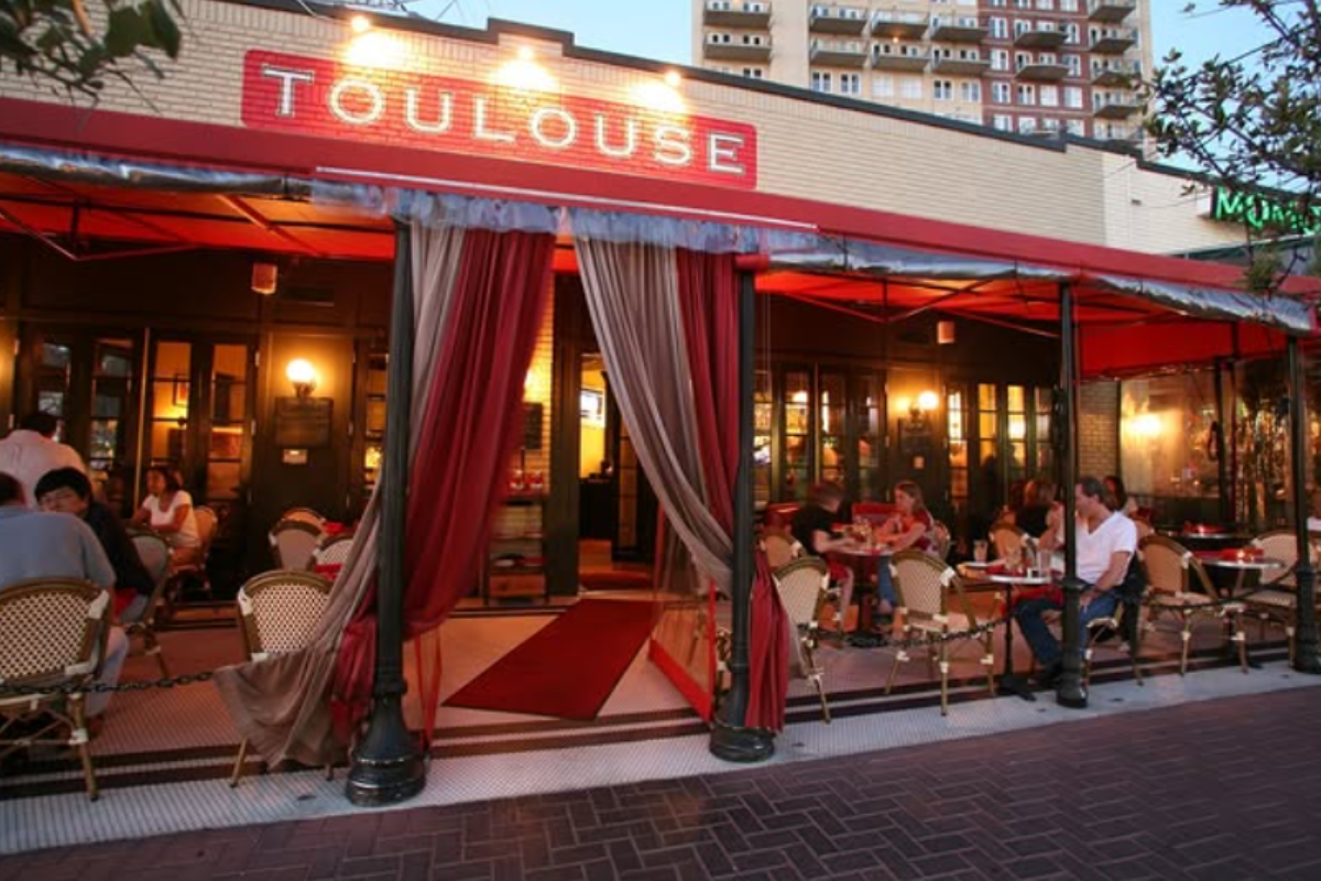 Toulouse Cafe outdoor dining with red awning, draped entrance, wicker chairs, and cozy atmosphere as guests enjoy meals under warm evening lights.