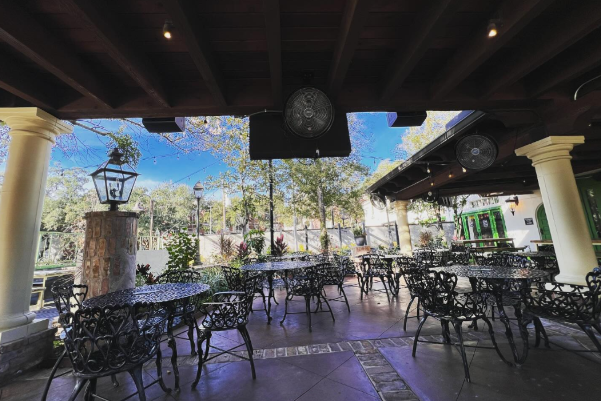 Verde Garden patio with black wrought iron tables and chairs, string lights, stone columns, fans, and surrounding greenery under a bright blue sky.