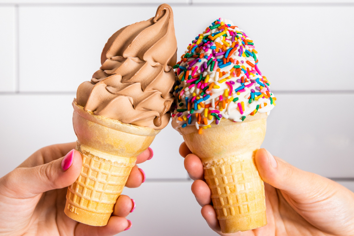 Two hands holding Carvel vanilla soft serve with rainbow sprinkles and chocolate soft serve in cake cones against a white tiled background at Buttermilk Baby.