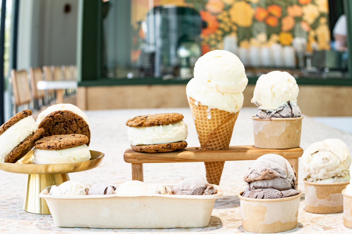 Assorted Honeychild’s Sweet Cream scoops, cones, sandwiches, and flights displayed on a table with a colorful floral mural in the background.
