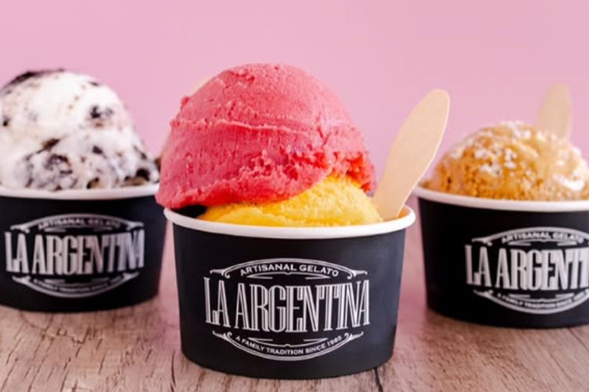 Three cups of La Argentina artisanal gelato with scoops of bright red, yellow, white, and brown flavors, served in black cups with wooden spoons.