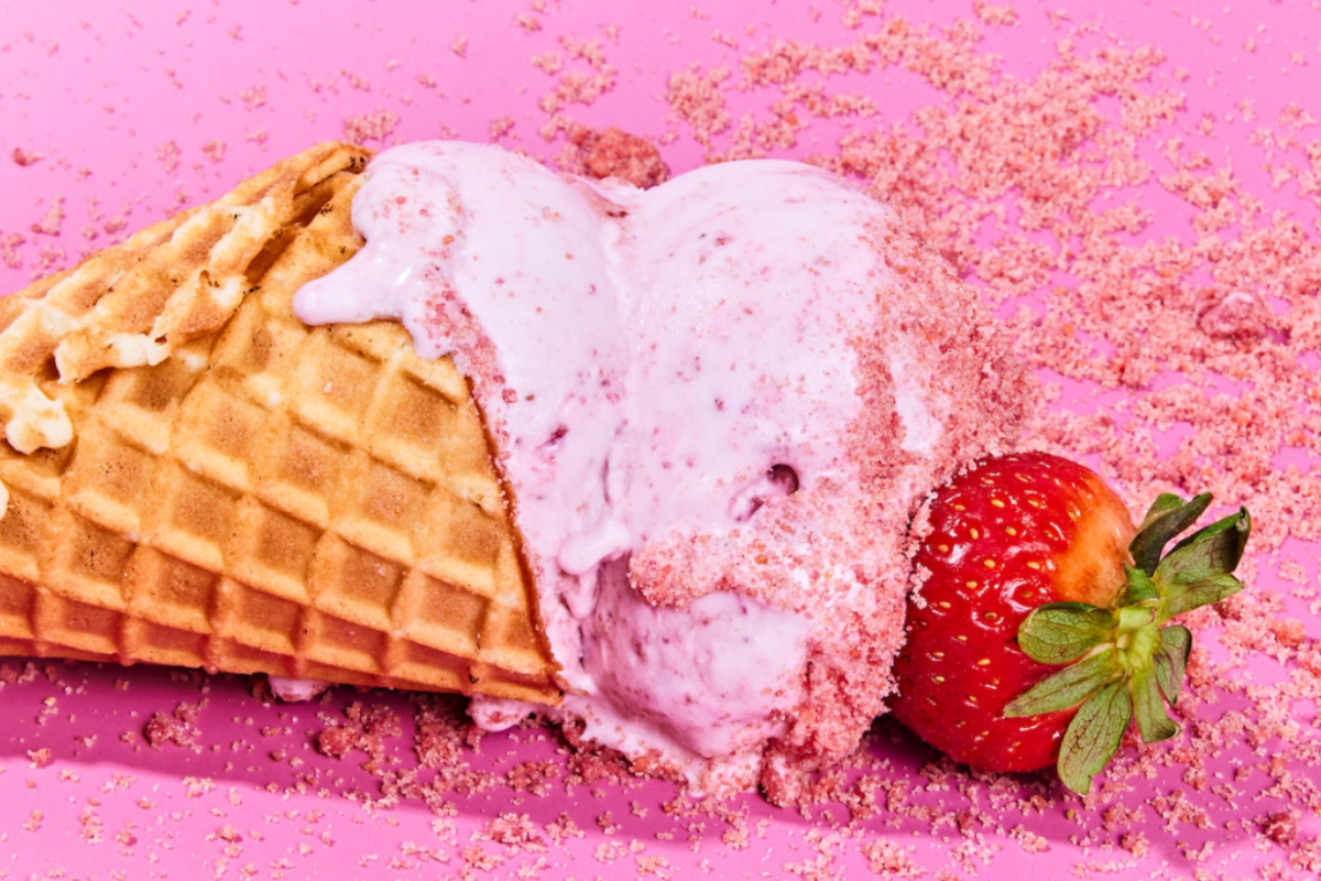 Waffle cone with Lick Honest strawberry ice cream melting on a pink surface, topped with pink crumbs and a fresh strawberry beside it.